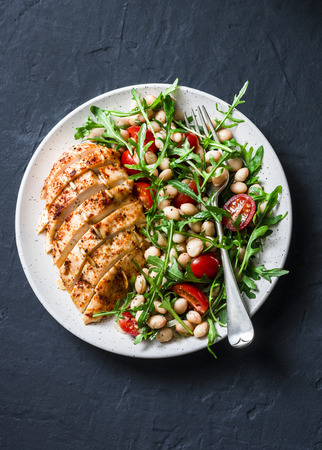 Delicious healthy lunch - baked chicken breast and white beans, arugula, cherry tomatoes salad on a dark background, top viewの写真素材