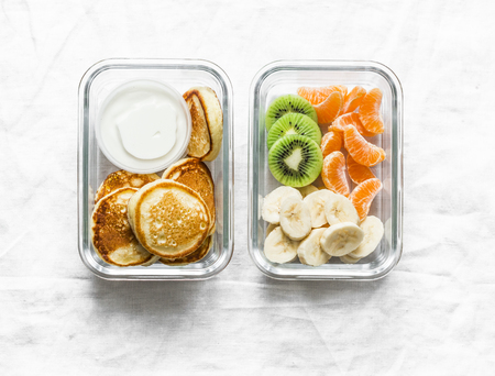 Healthy kids school lunch box - pancakes with sour cream and banana, kiwi, tangerine fruit. Delicious snack on a light background, top viewの写真素材