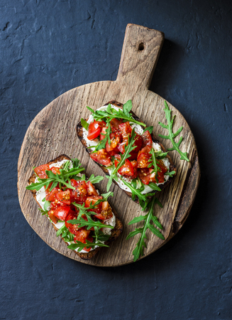 Tomatoes and rocket salad whole grain bread bruschetta on a wooden chopping board on a dark background, top view. Copy spaceの写真素材