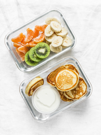 Healthy kids school lunch box - pancakes with sour cream and banana, kiwi, tangerine fruit. Delicious snack on a light background, top viewの写真素材
