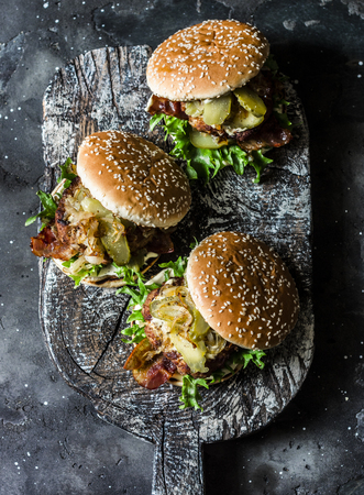 Classic homemade burger with pork cutlet, bacon, pickles, fried onions and mayonnaise mustard sauce on wooden chopping board, on dark backgroundの写真素材