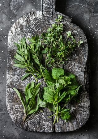 Aromatic garden herbs, food ingredients, seasonings - sage, thyme, mint, tarragon on a wooden rustic chopping board on a dark background, top viewの写真素材