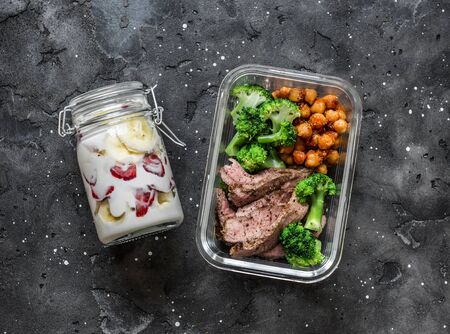 Balanced lunch box - breakfast - greek yogurt with strawberries and banana and lunch - steak, spicy chickpeas and broccoli on a dark background, top viewの写真素材