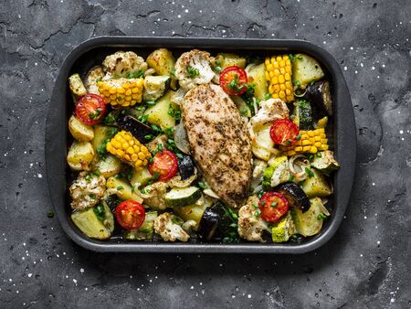 Chicken breast baked with potatoes, cauliflower, corn, eggplant, zucchini, cherry tomatoes on a baking sheet on a dark background, top view. Healthy food conceptの写真素材