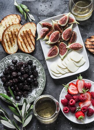 Mediterranean style snack appetizers - dried olives, figs, cheese, grilled bread, strawberries, raspberries and white wine on a dark background, top viewの写真素材