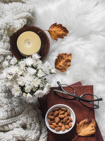 Cozy home interior - stack of books, almond nuts, knitted plaid, candle, chrysanthemum bouquet, fur carpet on a light background, top viewの写真素材