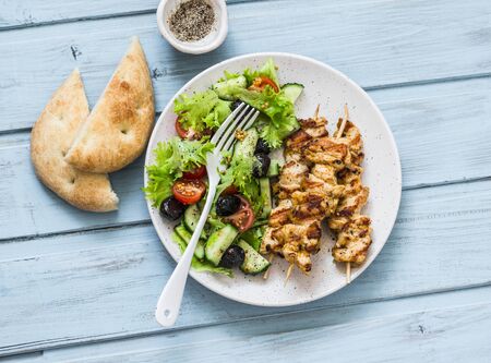 Chicken souvlaki kebabs, sun dried olives, tomatoes, cucumbers green salad and focaccia on blue background, top viewの写真素材