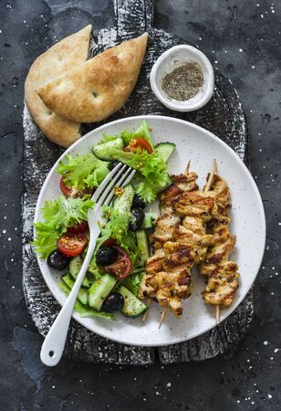 Chicken souvlaki kebabs, sun dried olives, tomatoes, cucumbers green salad and focaccia tortilla on dark background, top viewの写真素材