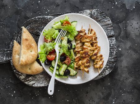 Chicken souvlaki kebabs, sun dried olives, tomatoes, cucumbers green salad and focaccia tortilla on dark background, top viewの写真素材