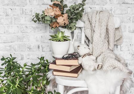 Concept of a cozy home. Old chair, stack of books, knitted plaid, teddy bear toy, succulent flower, dry eucalyptus's branches on the background of a light wallの写真素材