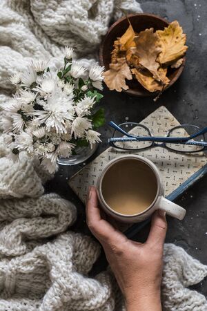 Woman's hand holding coffee mug, book, cozy plaid, autumn leaves on dark background, top viewの写真素材