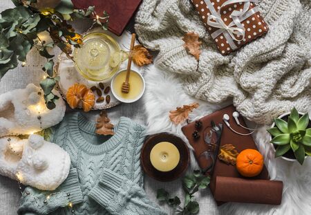 Cozy winter autumn things. Home coziness still life - soft slippers, knitted sweater, plaid, green tea with honey, tangerines, dry leaves, books, garland, gift box on a light background, top view. Autumn and winter relax mood. Flat layの写真素材