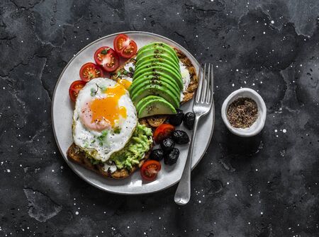 Delicious brunch - fried egg, avocado, grilled bread, dried olives, cherry tomatoes. Delicious healthy breakfast, snack on a dark background, top viewの写真素材
