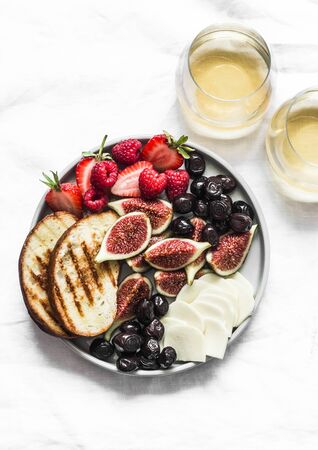 Glasses of white wine and snack appetizers tapas plate with cheese, figs, olives, berries on a light background, top viewの写真素材