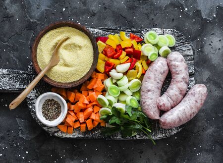 Ingredients for cooking lunch - chopped vegetables, polenta, sausages on a rustic cutting board on a dark background, top view. Making polenta with sausage vegetables stewの写真素材