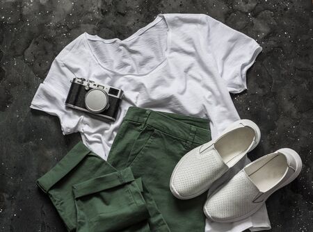 Women's clothing for summer spring walks - cotton green trousers, a basic white t-shirt, white sneakers and a vintage camera on a dark background, top view. Fashion conceptの写真素材