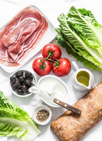 Ingredients for bruschetta - prosciutto, ciabatta bread, romano salad, olives, tomatoes, cream cheese on a light background, top view. Flat layの写真素材