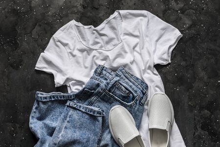 Simple women's look - white t-shirt, mom's jeans, white sneakers on a dark background, top view. Fashion concept, flat lay. Clothes for walkingの写真素材