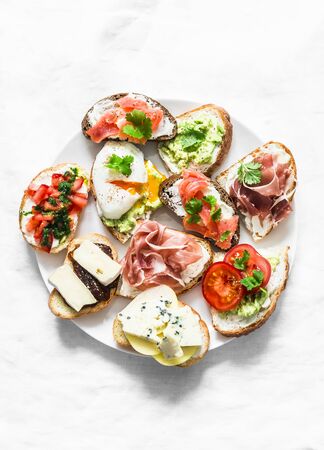 Variety of tapas sandwiches plate - sandwiches with prosciutto, avocado, salmon, egg, tomatoes, jamon, gorgonzola, brie, pear on a light background, top view. Delicious snack, appetizers. Copy spaceの写真素材