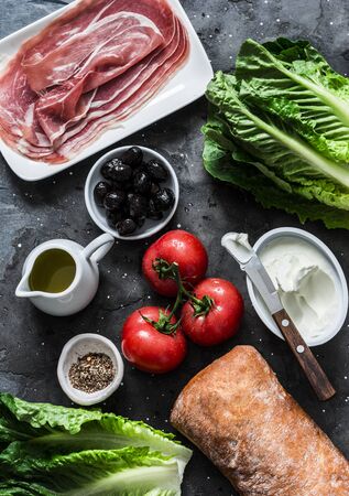 Ingredients for bruschetta - prosciutto, ciabatta bread, romano salad, olives, tomatoes, cream cheese on a dark background, top view. Flat layの写真素材