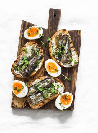 Delicious appetizer, tapas - soft boiled eggs and canned sardines sandwiches on a cutting board on a light background, top viewの写真素材