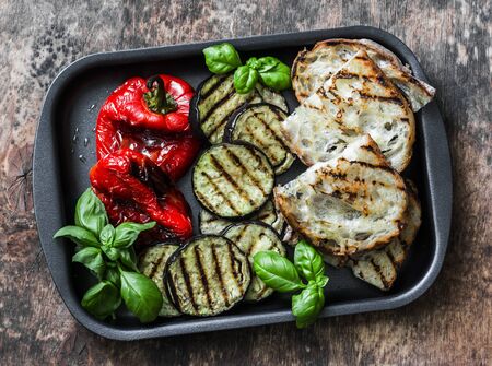 Summer grilled vegetables - eggplant, bell peppers and ciabatta bread, basil in baking sheet on wooden background, top viewの写真素材