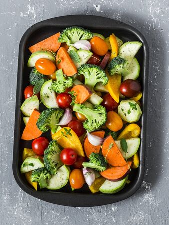 Cooking lunch. Raw fresh vegetables on a baking sheet. Sweet potato, zucchini, sweet pepper, cherry tomatoes, garlic, broccoli cabbage - food ingredients  on grey background, top viewの写真素材