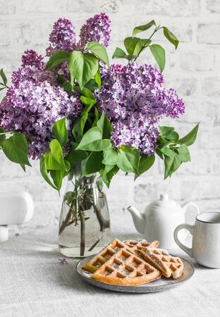 Cozy still life - waffles, lilac bouquet, teapot on the table in a bright roomの写真素材