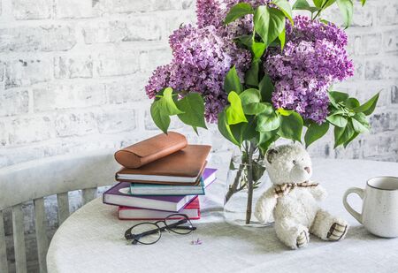 Cozy home still life - bouquet of lilacs, stack of books, glasses, toy bear, mug on the table in a bright roomの写真素材