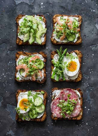 Variety breakfast, appetizer, tapas whole grain bread sandwiches with cream cheese, egg, asparagus, avocado, cucumber, shrimp, salami, micro greens on a dark background, top viewの写真素材
