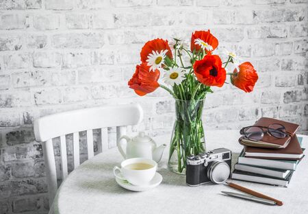 Bunch of poppies, stack of books, cup of green tea, vintage camera on a table in a bright room. Cozy home interior conceptの写真素材