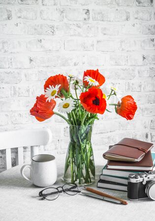 Bouquet of poppies, daisies, stack of books, cup of tea, camera on a table in a bright room. Cozy interior conceptの写真素材