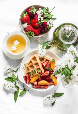 Green herbal tea, waffles with strawberries and apricots, flowers, strawberry bowl on a light background, top view. Flat layの写真素材