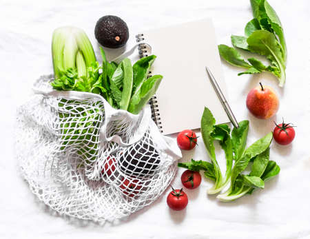 Grocery food concept. Fresh celery, green salad, avocado, cherry tomatoes in a rag eco bag and blank planning notepad on a light background, top viewの写真素材