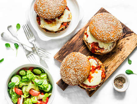 Homemade whole grain bun, grilled chicken, tomato sauce, mozzarella cheese burgers on the cutting board and tomato, cucumber, avocado salad on a light background, top view. Flat layの写真素材