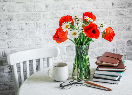 Cozy interior home concept. Bouquet of poppies, daisies, stack of books, cup of tea on a table in a light roomの写真素材