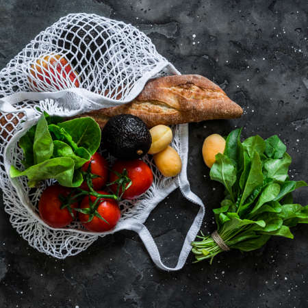 Fresh baguette bread, ripe tomatoes, apricots, spinach, avocado in a textile eco bag on a dark background, top view. Grocery conceptの写真素材