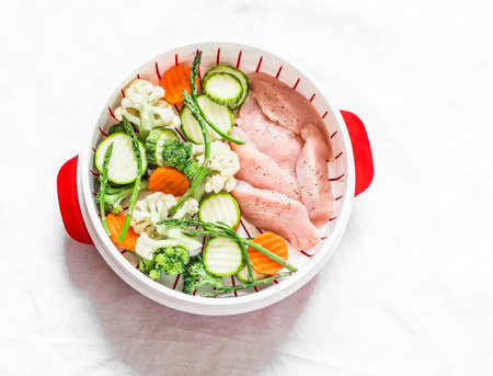 Cooking a healthy balanced meal concept. Fresh vegetables broccoli, zucchini, carrots, asparagus and chicken fillet in a steamer on a light background, top viewの写真素材