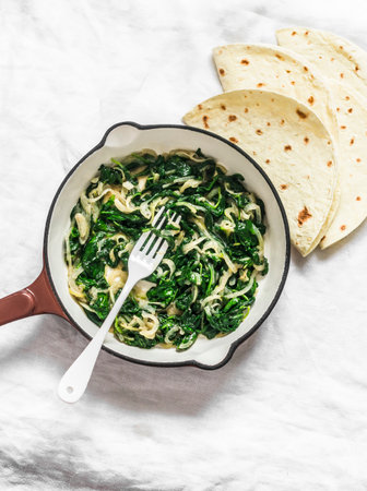 Stewed spinach in a cooking pan and tortillas on a light background, top view