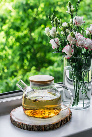 Green herbal tea in a glass teapot and a bouquet of flowers on the windowsill in the apartment opposite the green summer gardenの写真素材