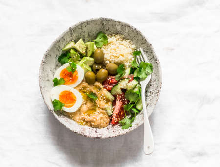 Delicious healthy vegetarian breakfast, brunch, lunch - couscous, boiled egg, avocado, fresh vegetables, hummus in one bowl on a light background, top viewの写真素材