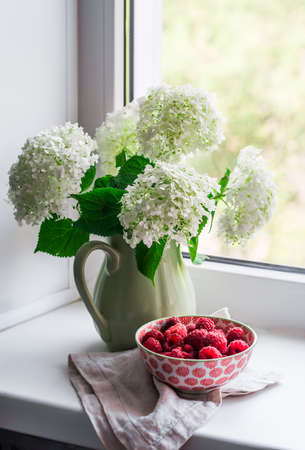 Bouquet of white hydrangeas in a vintage ceramic jug and bowl with raspberry on the windowsill. Home interior decorの写真素材