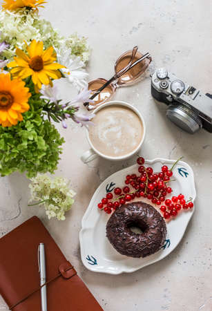 Delicious brunch - donut with chocolate icing, cappuccino, bouquet of flowers, camera, planner on a light stone background, top viewの写真素材