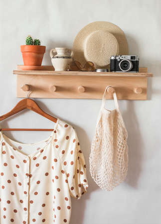 Women's summer blouse with polka dots on a hanger on a wooden shelf with a straw hat, an eco bag, a camera in a bright room. Vacation moodの写真素材