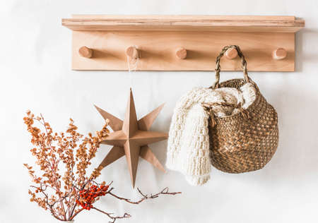 A wooden hanger with a scandinavian handmade christmas star, a knitted scarf in a jute basket and dried flowers on a light wall in the hallwayの写真素材