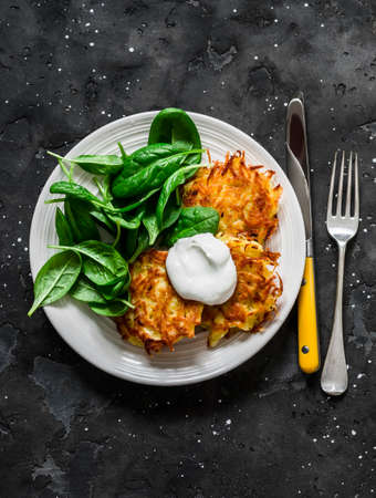 Potato latkes with sour cream and spinach salad on a dark background, top view