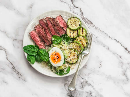 Balanced delicious healthy lunch - medium roast beef steak, grilled zucchini and couscous, avocado, spinach, egg salad on a light background, top viewの写真素材