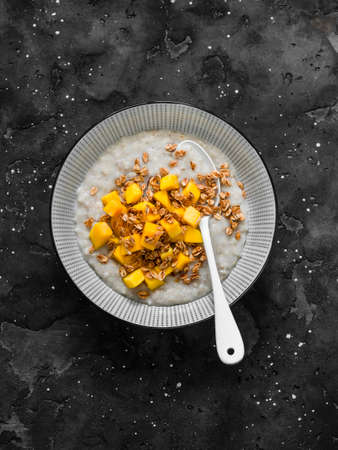 Oatmeal coconut milk gluten free porridge with fresh mango and crispy granola on a dark background,...