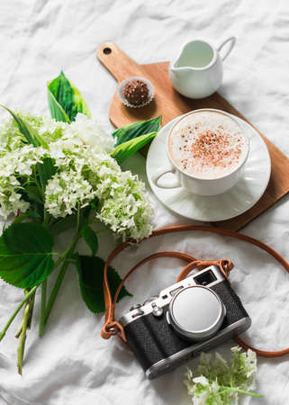 Cozy home still life - cappuccino in bed, bouquet of flowers and retro camera on a light background,...
