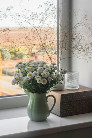 A bouquet of chrysanthemums in a vintage ceramic jug, a craft box, a candle on the windowsill with an autumn view outside the window. Cozy home interiorの写真素材